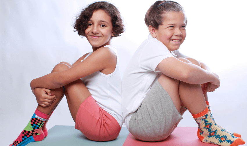 Two smiling children sitting back-to-back wearing vibrant and colourful socks, promoting Intots's inclusive designs. Highlighting Purpl's exclusive disabled discounts, making accessible fashion and adaptive clothing affordable for families in the UK.