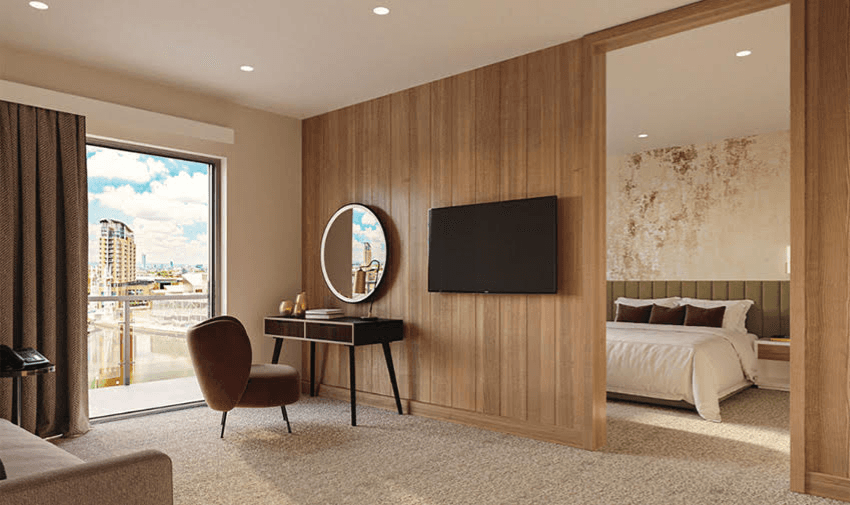 A modern hotel room featuring a wooden accent wall with a mounted flat-screen TV. A round mirror and chair sit on a desk next to a sliding glass door opening to a balcony with city views. A cosy bedroom is visible through an open doorway.