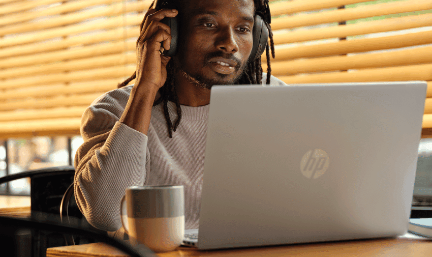 A person with long hair sits at a table in front of an HP laptop, wearing headphones, and appears focused. A mug is on the table beside the laptop. Blinds cover the window behind them, allowing some sunlight to filter through.