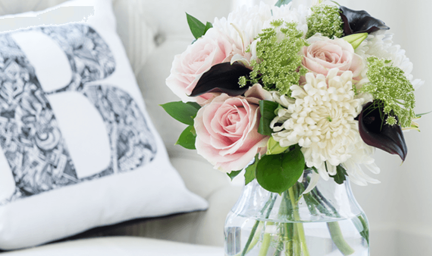 A clear glass vase holds a bouquet of soft pink roses, white chrysanthemums, and dark calla lilies. It's placed on a white cushion chair with a patterned pillow featuring the letter "B" in the background.