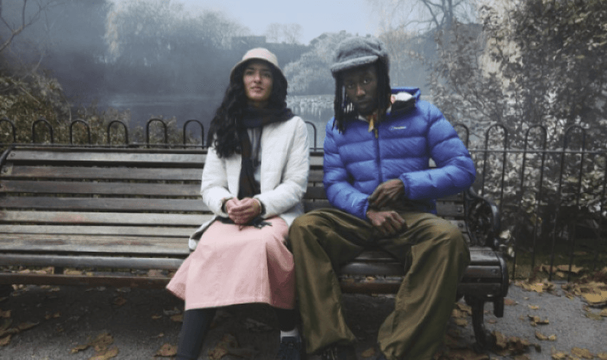 A woman and a man sit on a park bench. The woman wears a white jacket, pink skirt, and hat. The man wears a blue puffer jacket, green pants, and a cap. Fallen leaves are scattered on the ground, with trees and a body of water in the background.