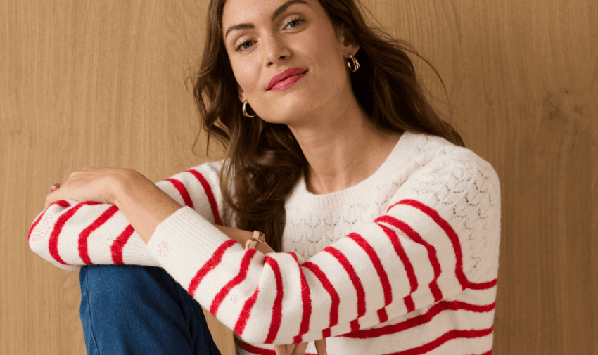 A woman with long brown hair sitting casually, wearing a white sweater with red stripes and blue jeans. She is smiling slightly against a wooden background. Disabled clothing discounts, adaptive fashion deals UK, inclusive offers for disabled shoppers.