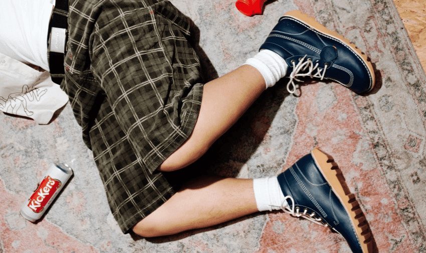 A person wearing plaid shorts, white socks, and dark blue shoes with tan soles is lying on a rug. A can labeled "Kickers" is nearby on the rug.