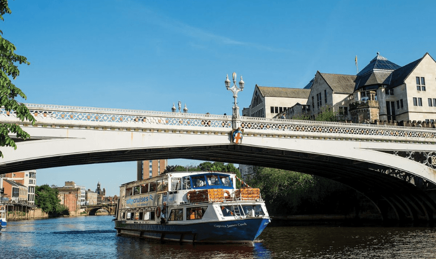 A sightseeing boat cruises along a river under an ornate bridge on a clear day. In the background, there are historic buildings and lush trees lining the riverbank. People can be seen walking across the bridge.