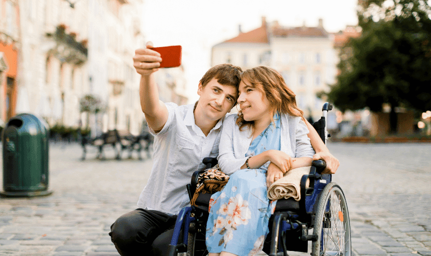 A young man and woman are taking a selfie outdoors. The woman is sitting in a wheelchair, and both are smiling. The man is holding a phone, capturing the photo. They are in a cobblestone square with blurred buildings and trees in the background.