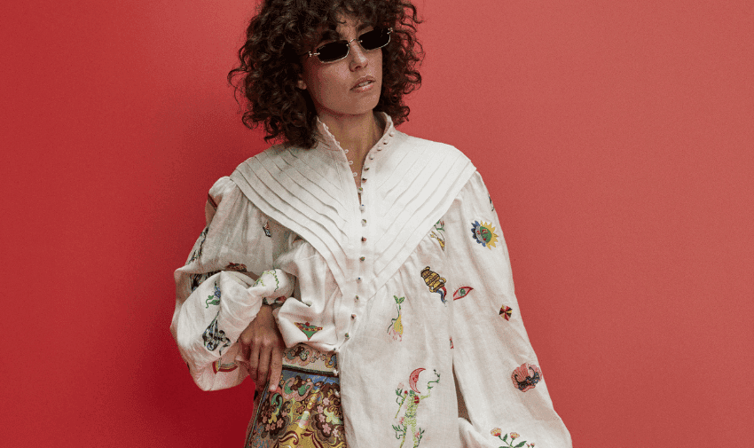 A person with curly hair wearing small, rectangular sunglasses poses against a red background. They are dressed in a white, buttoned-up shirt with colourful embroidery and pleated details, paired with a patterned skirt.