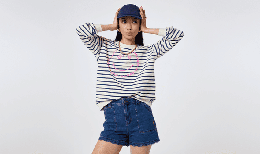 A young person strikes a playful pose while wearing a blue cap, a white and navy striped long-sleeve shirt with a pink graphic on the front, and blue denim shorts. The background is plain and light-coloured.