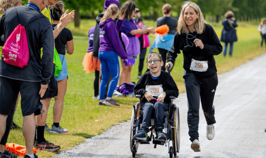 A woman joyfully running while pushing a young boy in a wheelchair, surrounded by cheering supporters at the Parallel Windsor race. Showcasing Purpl's disabled discounts for accessible and inclusive sports events in the UK.