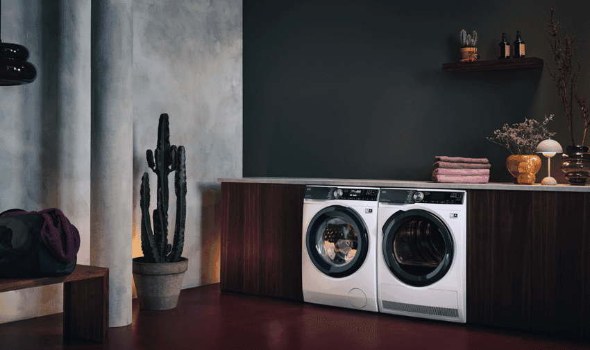 A modern laundry room features a front-loading washing machine and dryer side by side under a wooden countertop. The room is decorated with a potted cactus, a bench with a bag, and a small shelf with folded towels, plants, and bottles against a dark wall.