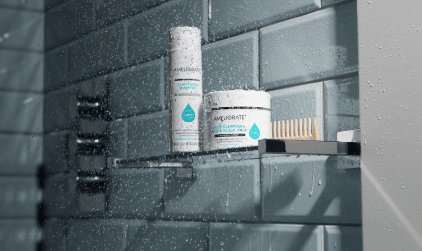 A shower shelf holding two white Ameliorate skincare products with blue labels and a beige brush. Water droplets cover the glass shelf and tiles, indicating the products are in use in a wet environment. The background features greyish-blue subway tiles.