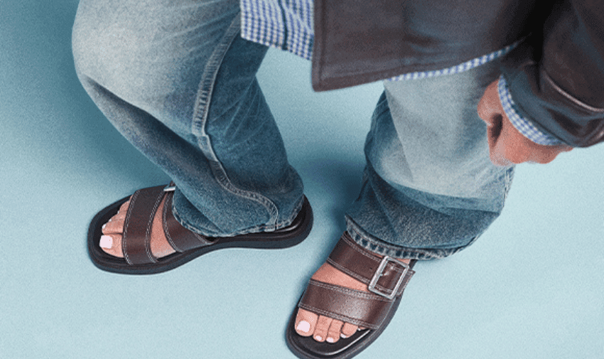 Person wearing a brown leather jacket, blue checkered shirt, and blue jeans stands on a light blue background. They are wearing brown leather sandals with a buckle, exposing their toes.
