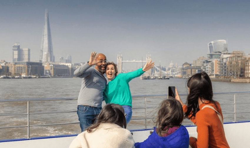 A group of people on a boat enjoying a sightseeing trip. Two adults are posing happily for a photo, with the Shard and Tower Bridge visible in the background. Three people, including a child, face the camera from the opposite side.