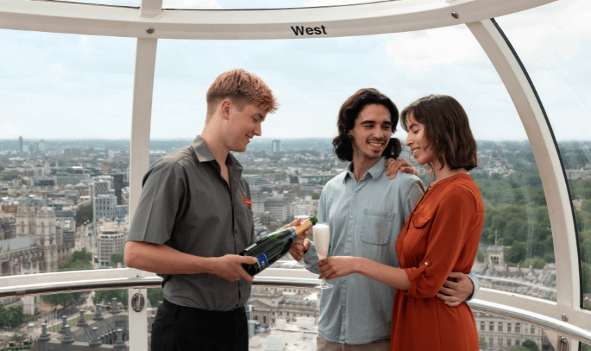 Couple enjoying a champagne experience inside a London Eye pod with a host serving drinks, set against a stunning panoramic view of London.