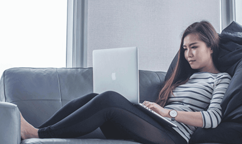 A woman with long hair sits on a grey couch, wearing a striped long-sleeve shirt and black leggings. She has a neutral expression and is focused on her open laptop, which is resting on her lap. She appears to be in a modern, well-lit room with large windows.