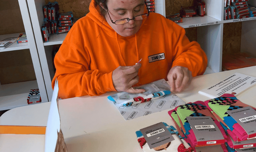 A person wearing an orange hoodie and glasses is putting tags on pairs of socks at a table. Various colourful socks with tags and boxes are scattered around the table. Shelves with more sock packages are seen in the background.