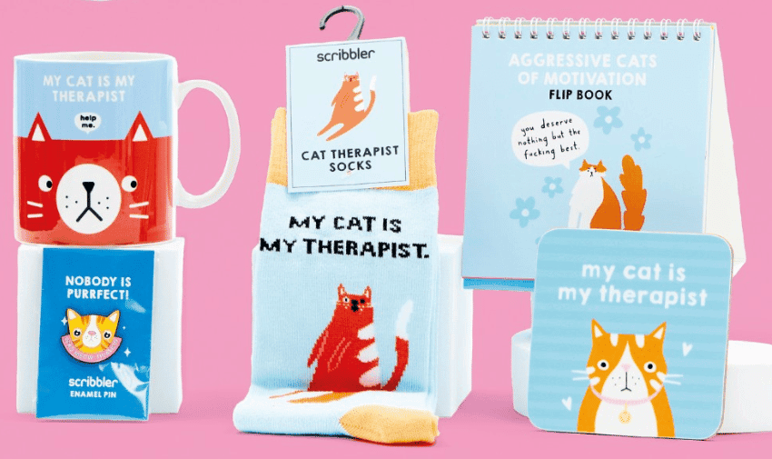 An assortment of cat-themed merchandise on a pink background. Items include a mug with "My Cat is My Therapist," a "Nobody is Purrfect" enamel pin, cat therapist socks, a flip book of motivational cats, and a notebook with "My cat is my therapist" on the cover.