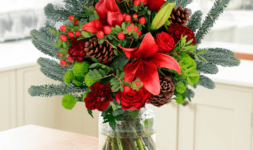 A festive bouquet of red lilies, roses, pinecones, and greenery arranged in a clear vase, set in a bright kitchen. Highlights shopping discounts for disabled people and Christmas gift ideas.