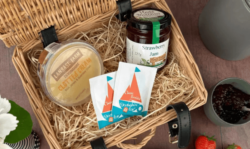 A wicker basket filled with a jar of strawberry jam, a pot of clotted cream and two tea sachets