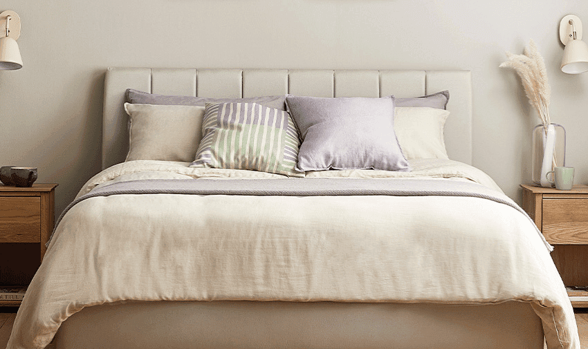 A cosy, well-made bed with layered bedding in neutral tones, adorned with several pillows in pastel shades and patterns. Wooden nightstands on each side hold minimal decor, including a lamp, a small plant, and a book. The wall behind is light-coloured with subtle decor.