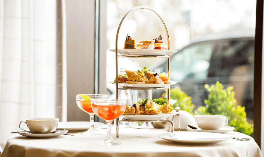 A three-tiered tray filled with various pastries, sandwiches, and desserts on a table set for afternoon tea. Two teacups and saucers, utensils, and two glasses of pink beverages are arranged beside the cake stand. The scene is by a window with natural light.