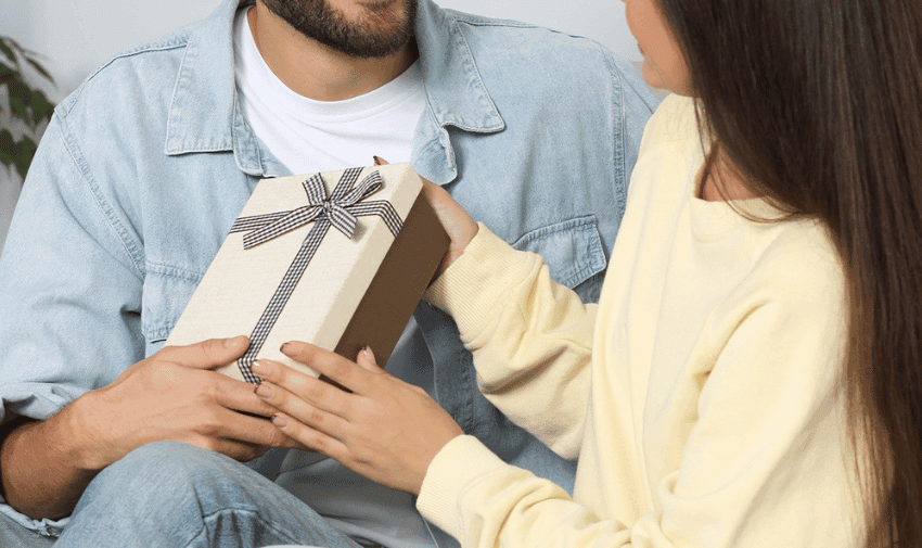 A person with long hair in a yellow sweater is handing a gift box with a ribbon to another person with a short beard wearing a denim jacket and white shirt. They appear to be indoors.
