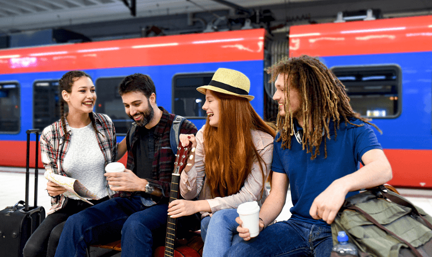 A group of young travellers sitting at a train station with luggage, enjoying coffee and a guitar while waiting for a red and blue train. Highlighting TrainPal and Purpl's exclusive disabled travel discounts, promoting affordable and accessible group travel options in the UK.