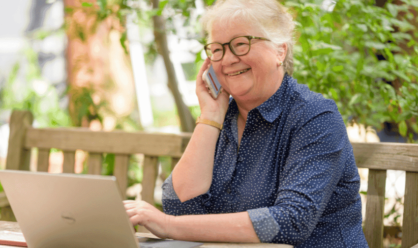 An older adult with short, white hair and glasses is sitting outdoors at a table, smiling while talking on a smartphone. They are wearing a blue, long-sleeved shirt with small white polka dots and are using a laptop.