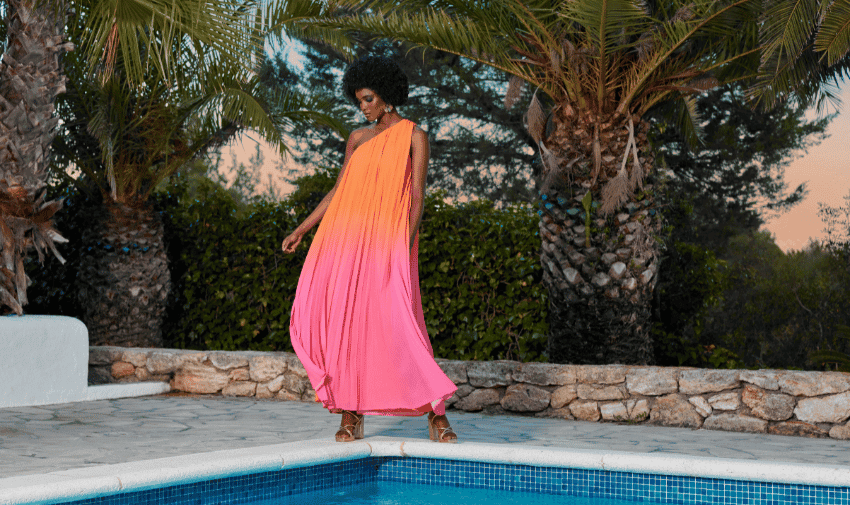 A woman in a flowing, gradient pink to orange dress stands near a pool, framed by palm trees and greenery. She looks down, her curly hair illuminated by the soft sunset light. The scene evokes a serene, tropical ambiance.