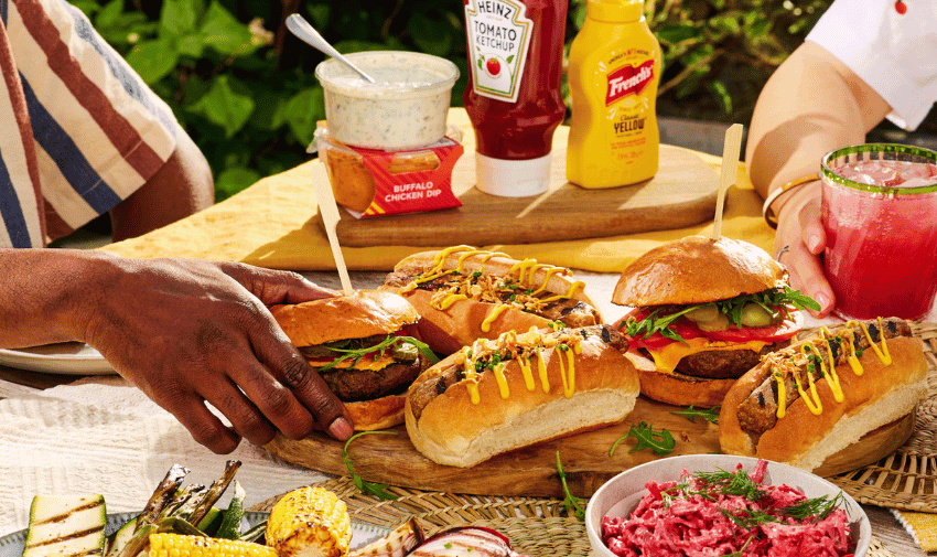 A colourful picnic spread on a wooden table includes sandwiches with grilled meats topped with mustard, fresh vegetables, corn on the cob, and a bowl of coleslaw. Condiments and a pink drink are visible in the background. Several hands are reaching for the food.