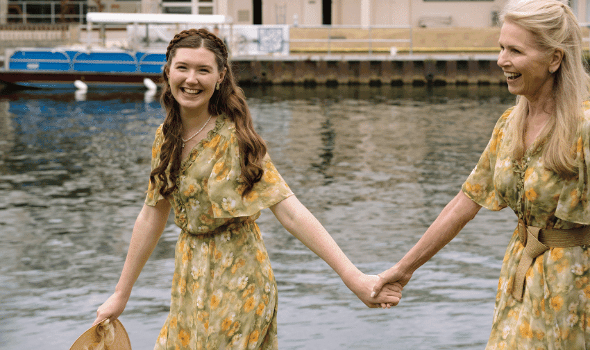 Two people, each wearing a green floral dress, walk hand in hand beside a body of water. The younger person carries a straw hat and has long curled hair, and the older person has long light hair and wears a brown belt. Both are smiling and appear joyful.