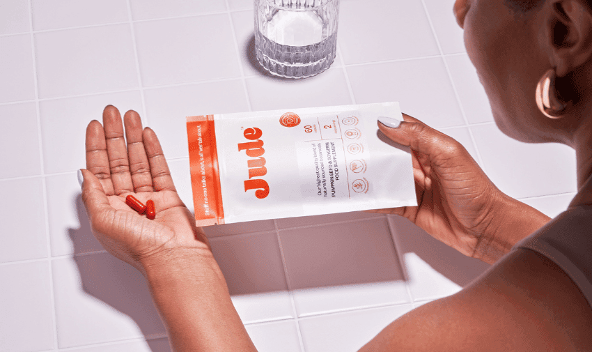 Image shows a lady looking at a packet of Jude daily support with two capsules in her hand and a glass of water nearby