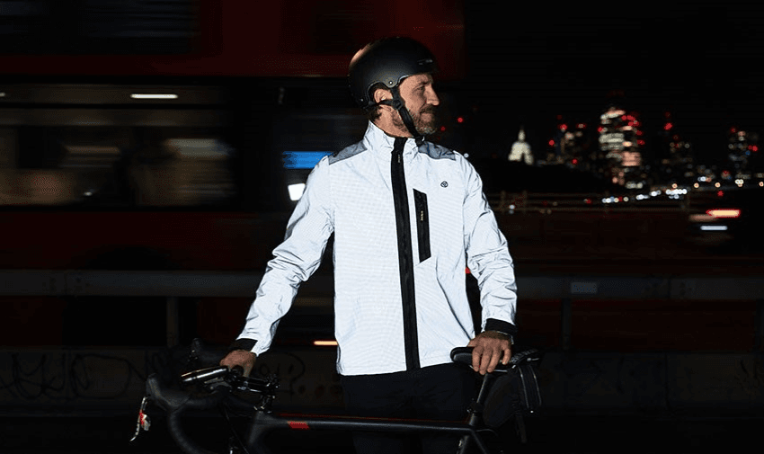 A cyclist standing with their bike, wearing a reflective Proviz jacket at night with a city skyline in the background. Showcasing Purpl's disabled discounts for stylish and safety-focused cycling apparel in the UK.