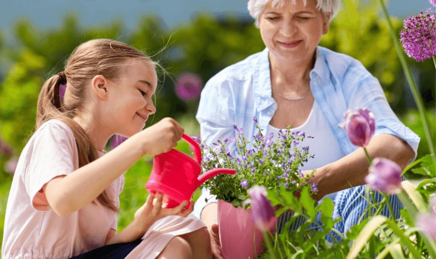 A young girl waters purple flowers with a pink watering can, while an elderly woman sits beside her, smiling, in a garden filled with blooming flowers and greenery.  https://trello.com/1/cards/670a6d01427bfbdd17a8ef4d/attachments/670a6dba5f344d966c3fb1a0/download/YouGarden_Purpl_Offer_Image.png