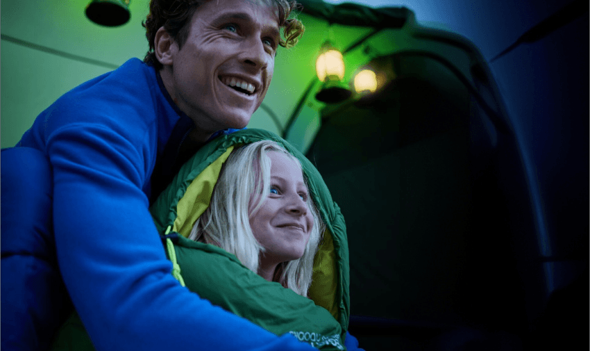 A man and a child are smiling and hugging inside a tent. They are wrapped in a warm sleeping bag, with lanterns glowing softly in the background. The scene conveys a cozy and joyful camping experience.