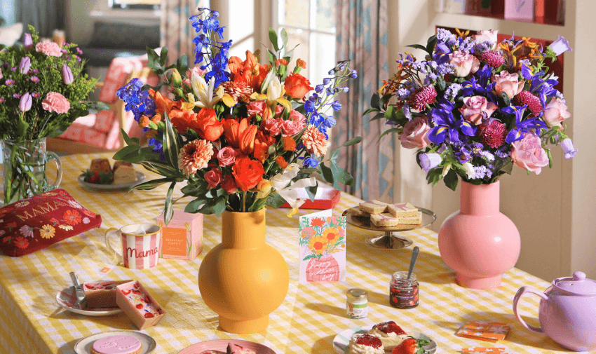 A colourful display of Bloom & Wild flower arrangements in vibrant vases on a dining table, surrounded by tea, jam, and treats, perfect for celebrations. Disabled discounts on flower delivery and home gifts.