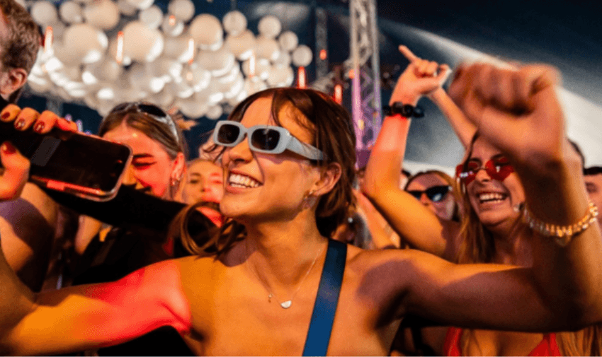 A woman enjoying a concert, smiling and dancing with her hands in the air, wearing fun 3D-style glasses in a vibrant crowd.