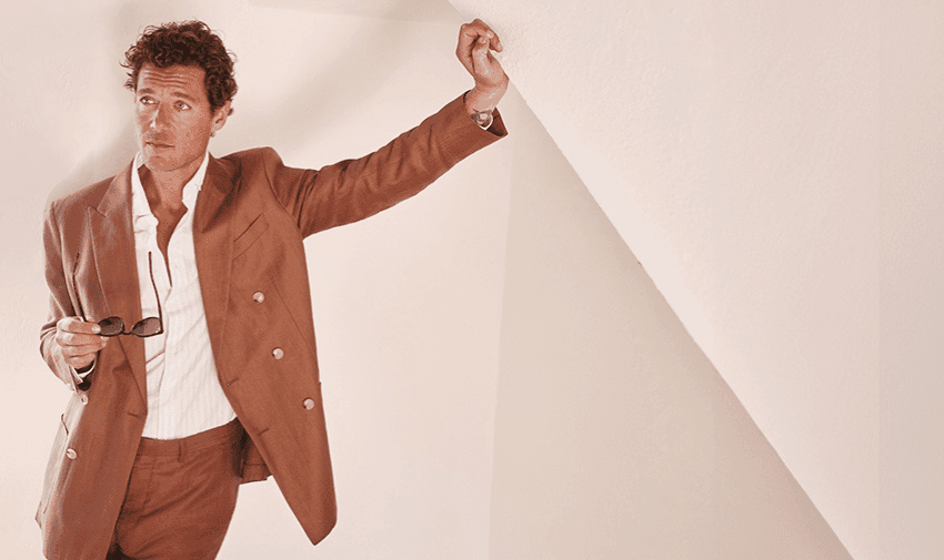 A man with curly hair is dressed in a brown suit and white shirt, holding sunglasses in one hand while leaning against a white wall with the other. He gazes off into the distance thoughtfully. The background is minimalistic and bright.