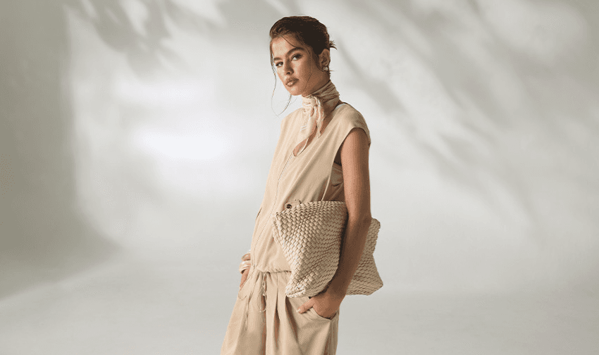 A woman dressed in a soft beige outfit with a matching woven clutch bag, standing against an elegant light background. Showcasing Purpl's disabled discounts for contemporary and accessible fashion from Boohoo in the UK.