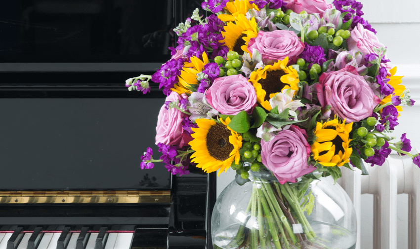 A vibrant bouquet of sunflowers, pink roses, and purple flowers in a clear vase sits on a piano next to a keyboard.