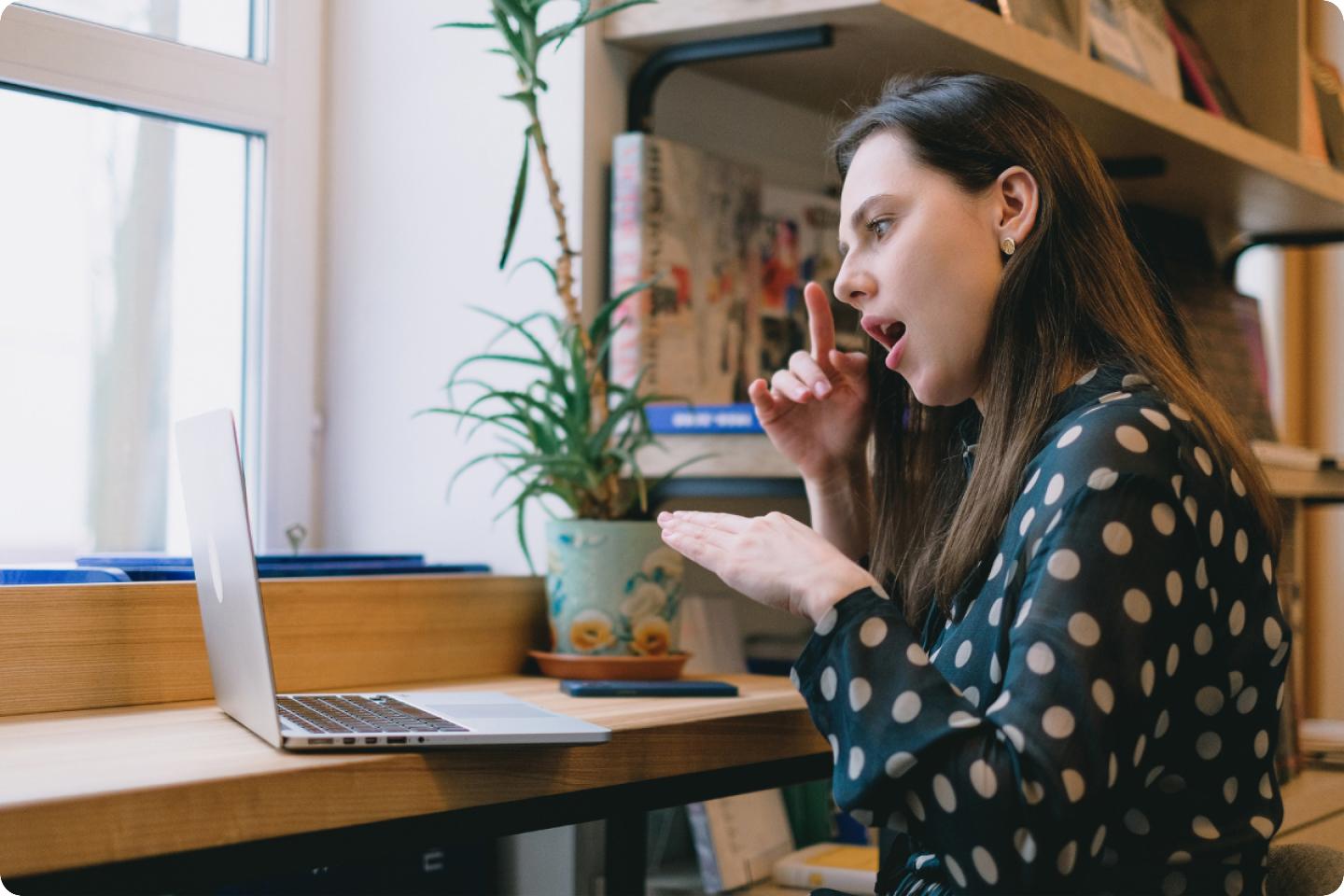 A woman with long hair wearing a polka-dot blouse is sitting at a wooden desk and using sign language while looking at a laptop screen. The desk is near a window with a plant in a pot and shelves filled with books above it.