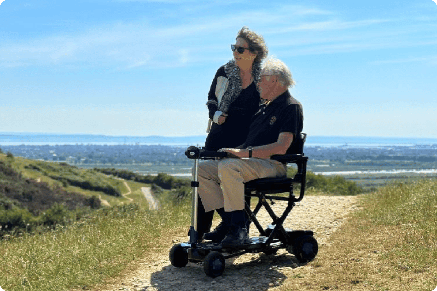 A woman stands next to an older man seated on a mobility scooter on a dirt path atop a hill. Both are dressed in casual clothes and sunglasses. The horizon shows a clear sky and a blur of distant landscape beneath them. They appear to be enjoying the scenery.