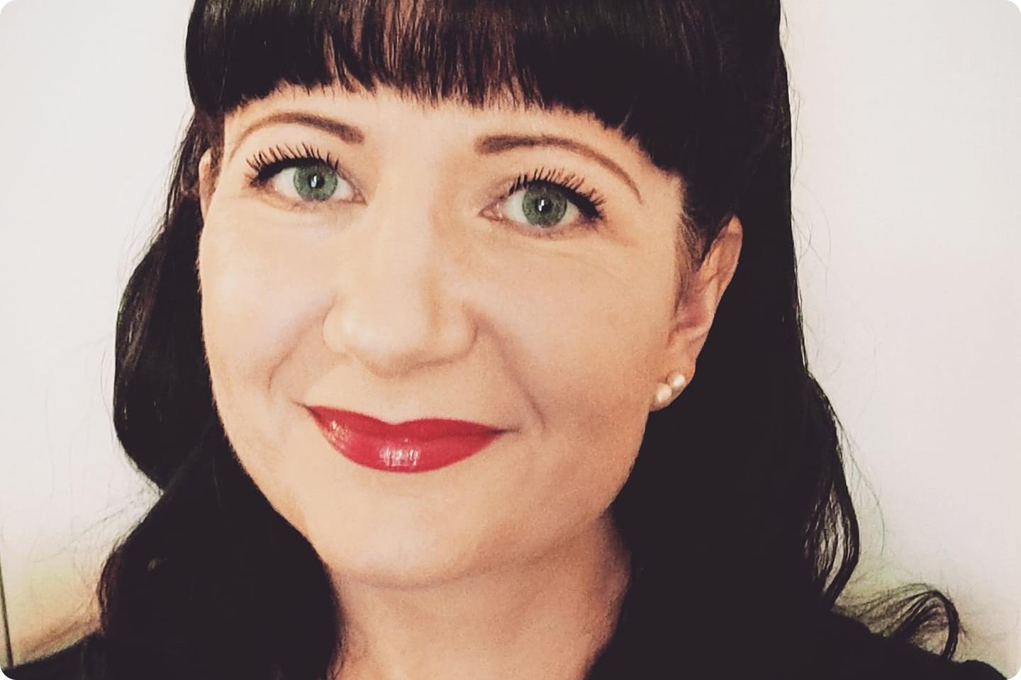 Carol, a woman with long dark hair, green eyes, and red lipstick smiles at the camera. She is wearing pearl earrings and has bangs covering her forehead. The background is plain and light-coloured.