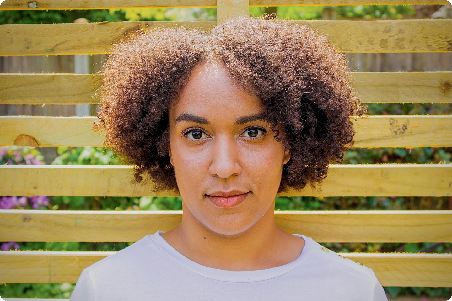 Image of Georgia Bondy standing in front of a panelled fence. She has brown skin and afro hair style and looks thoughtful.