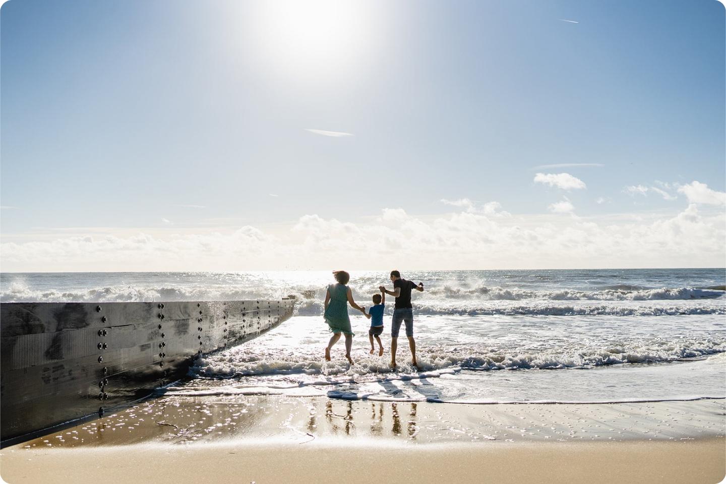 Photo shows 3 of Nicole’s family, dressed in summer clothes, playing near the edge of the ocean waves on a sunny beach. The parent on the left holds the child's hand in the middle, while the other parent stands to the right. The sky is partly cloudy and bright.