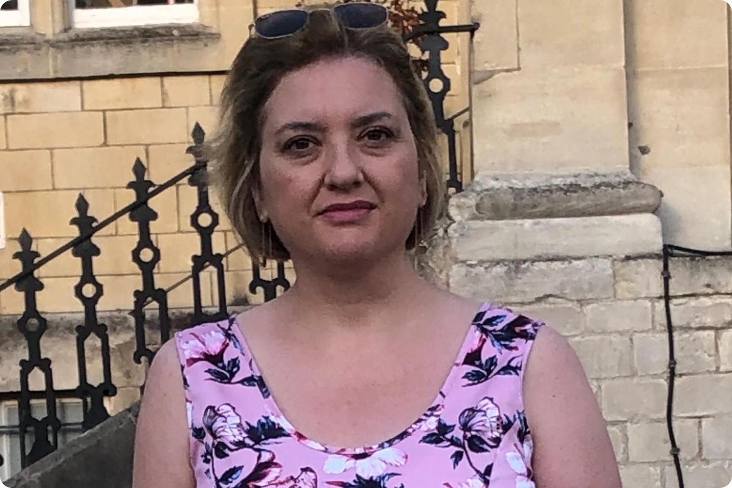 Photo of Paula a woman with short hair, wearing sunglasses on her head, stands in front of a beige stone building. She is dressed in a pink floral top and looks towards the camera with a neutral expression. Black decorative wrought iron fencing is visible in the background.