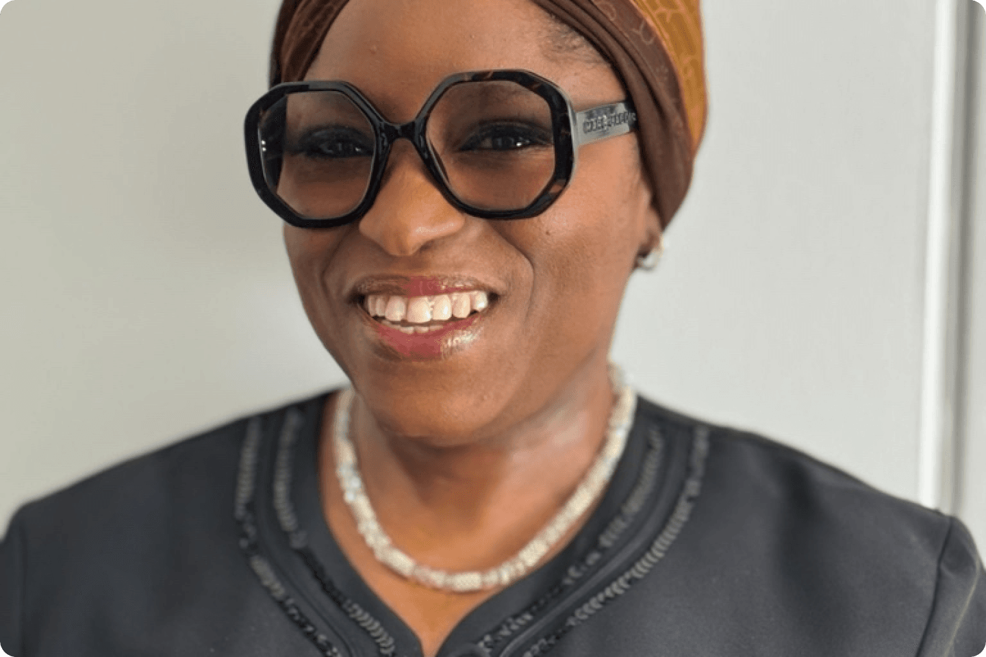 Funmi is smiling warmly, wearing large black-framed glasses, a patterned brown headscarf, and a dark top with a decorative neckline. A beaded necklace adorns their neck. The background is plain and light-coloured.