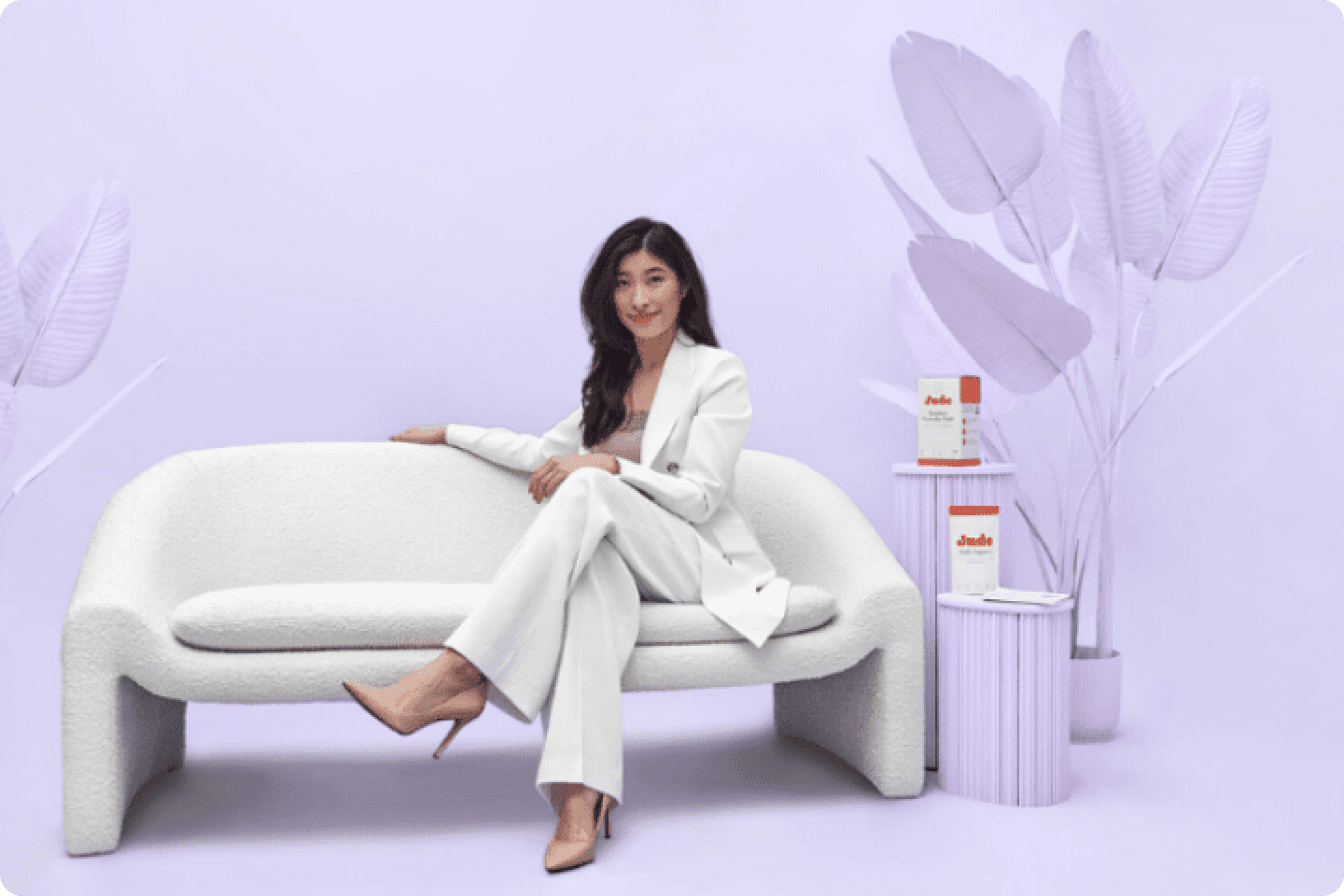 A woman in a white suit sits on a modern, white couch in a room with light purple walls and decor, including large, artificial leaves and two cylindrical side tables. There are two product boxes on one table. The scene has a minimalist, elegant feel.