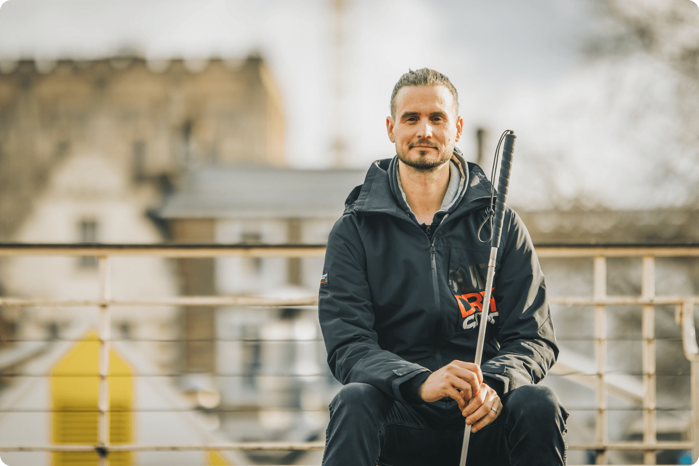 Rob Sears a man with a beard, wearing a black jacket, sits outdoors on a bench holding a white cane. The background features blurred urban architecture and a cloudy sky.