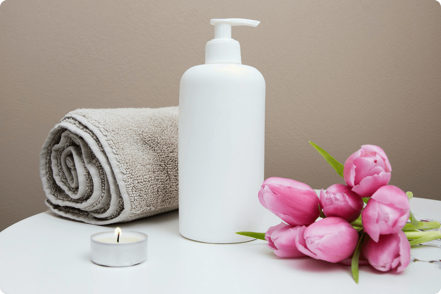 A white lotion bottle with a pump, a folded beige towel, a lit tea light candle, and a bunch of pink tulips are arranged on a white surface against a beige background. The scene portrays a calm, spa-like setting.