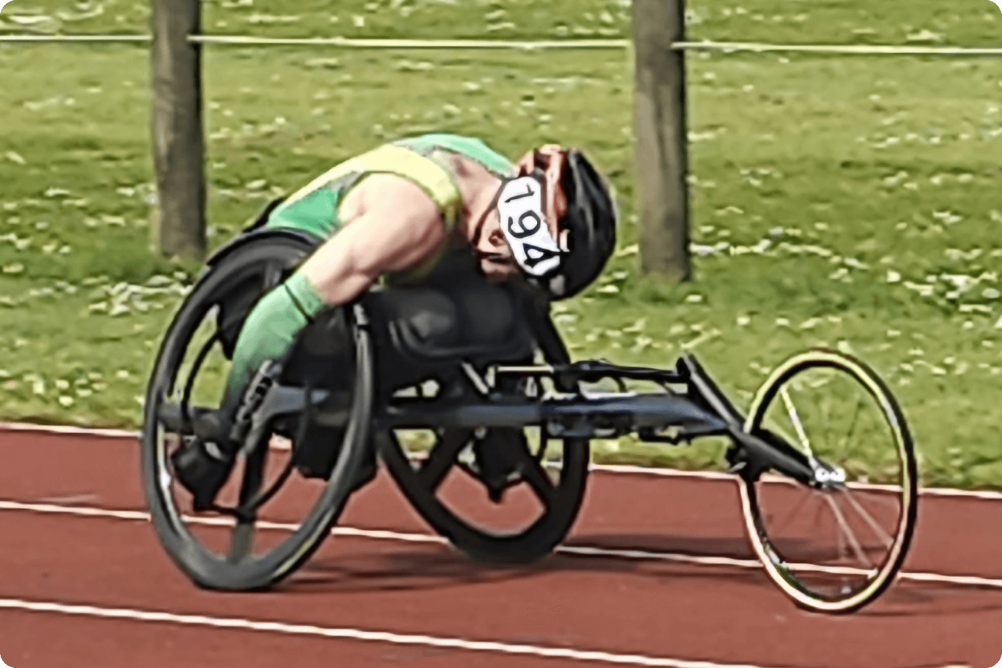 Toby Richardson in a sports wheelchair is on an outdoor racing track. The racer is wearing a helmet and a green outfit with the number 1964 on their head. They are in a forward-leaning position while pushing the chair. The background shows grass and fence posts.